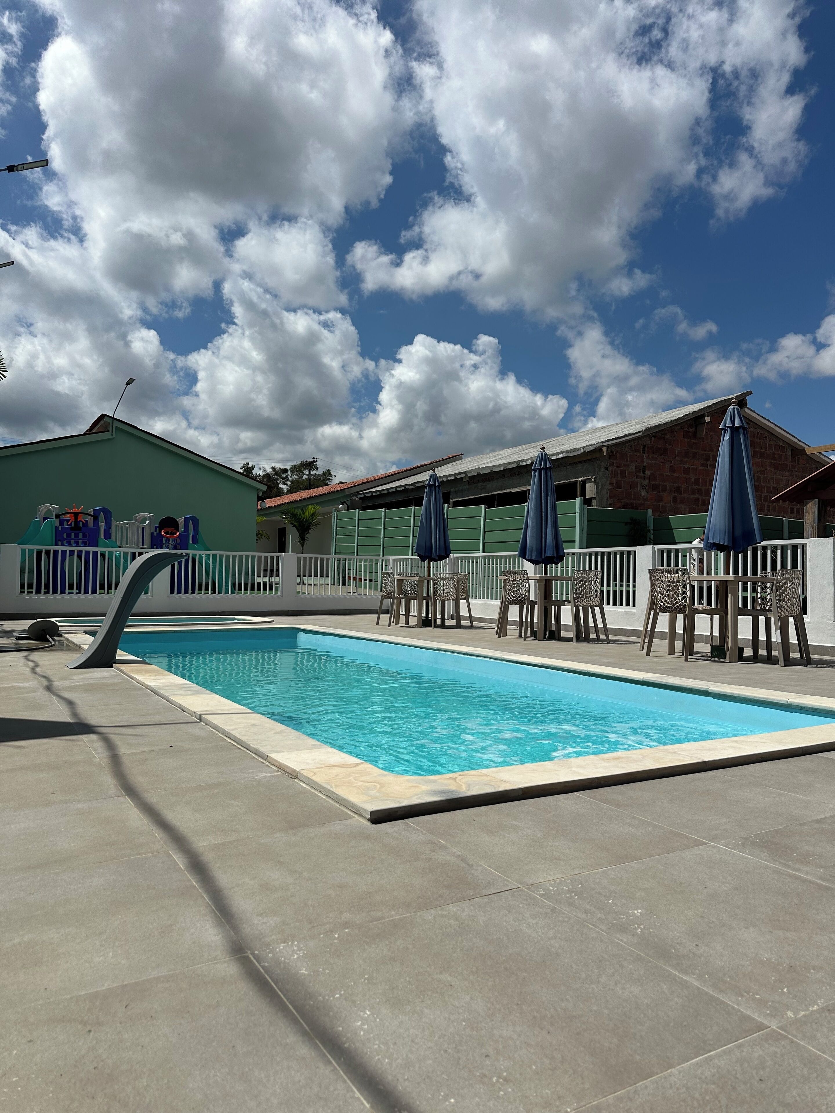 A swimming pool with a white fence, blue umbrellas, and a building with a green roof in the background.