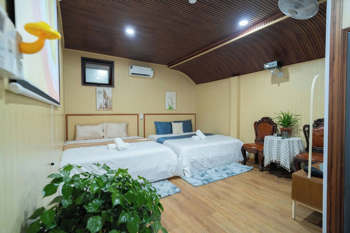 A hotel room with two beds, a ceiling fan, an air conditioning unit, a small table with a plant, and a wooden floor.