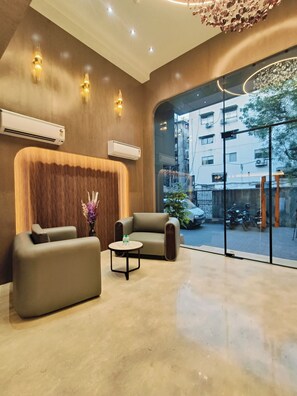 A modern lobby with two sofas, a small table with a vase of flowers, and a large window offering a view of the street. - Hotel Maskan Surat (Surat)