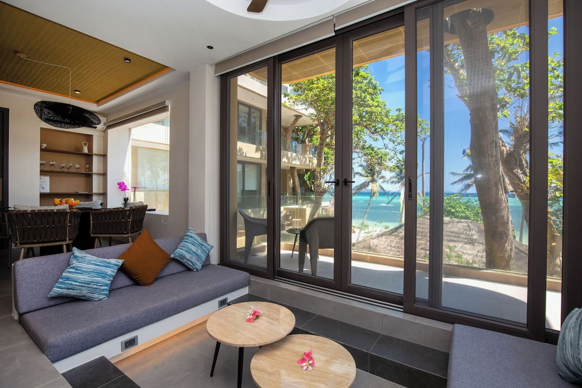 A modern living room with a sofa, two round tables, and a view of the beach and ocean through large sliding glass doors.
