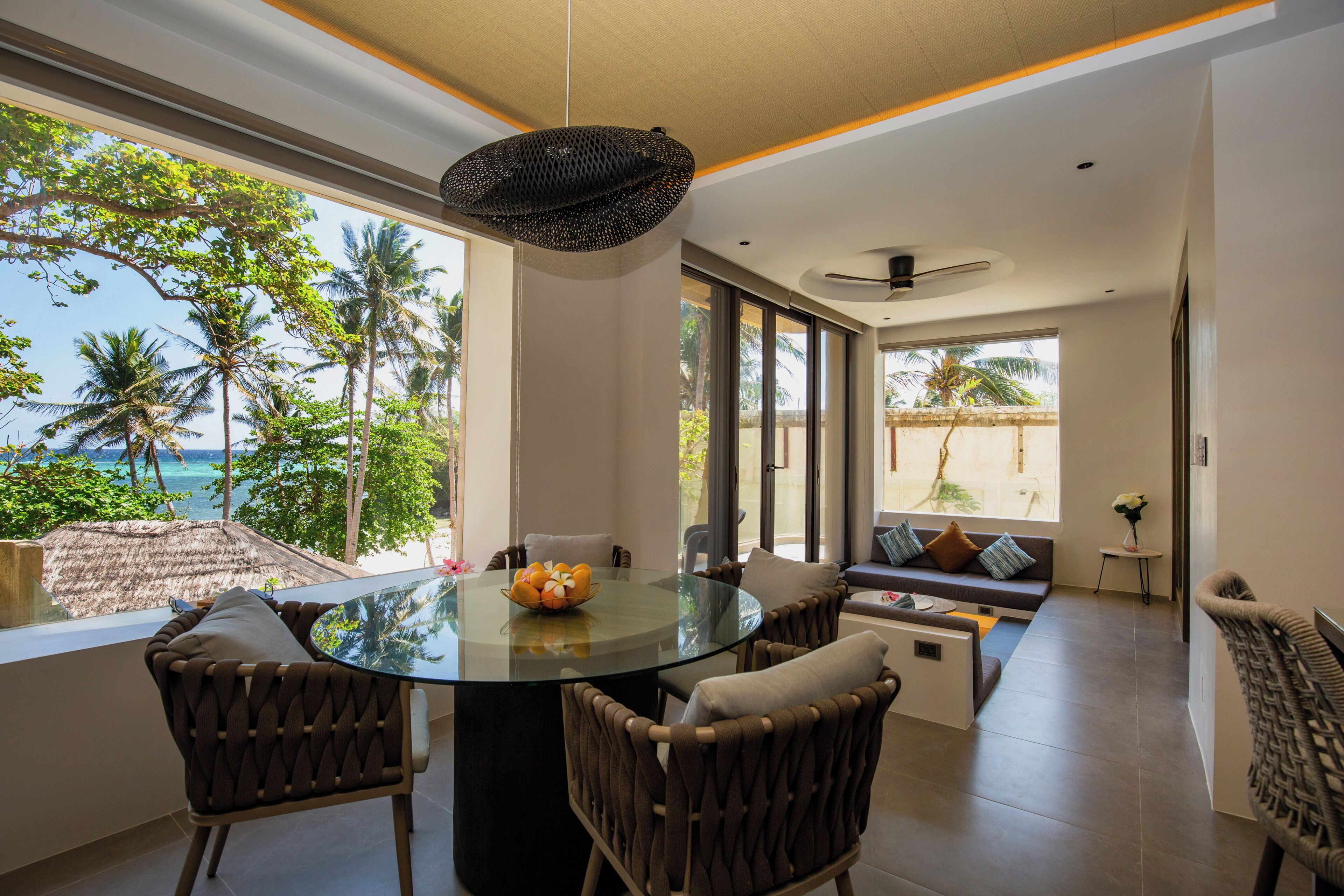 Un espace repas moderne avec une table en verre, des chaises en osier et une vue sur l’océan.