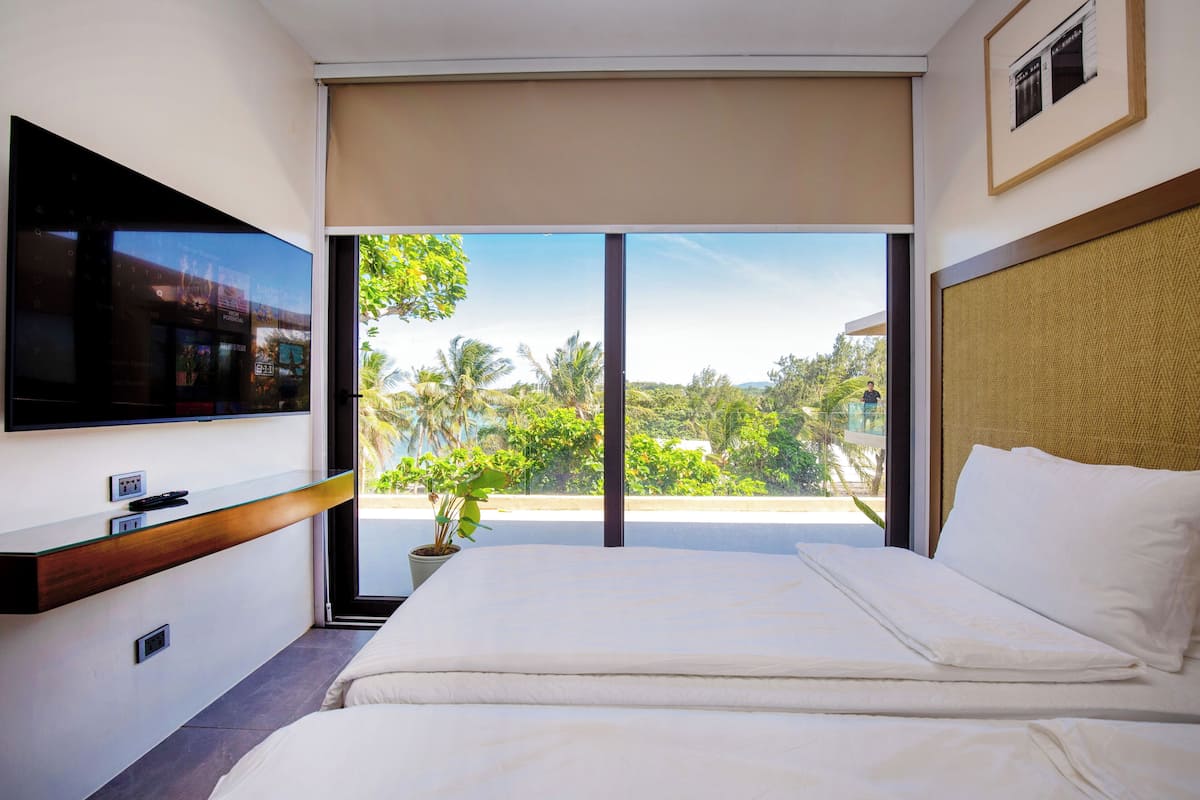 A hotel room with a bed, a TV mounted on the wall, a large window with a view of greenery, and a ceiling fan.