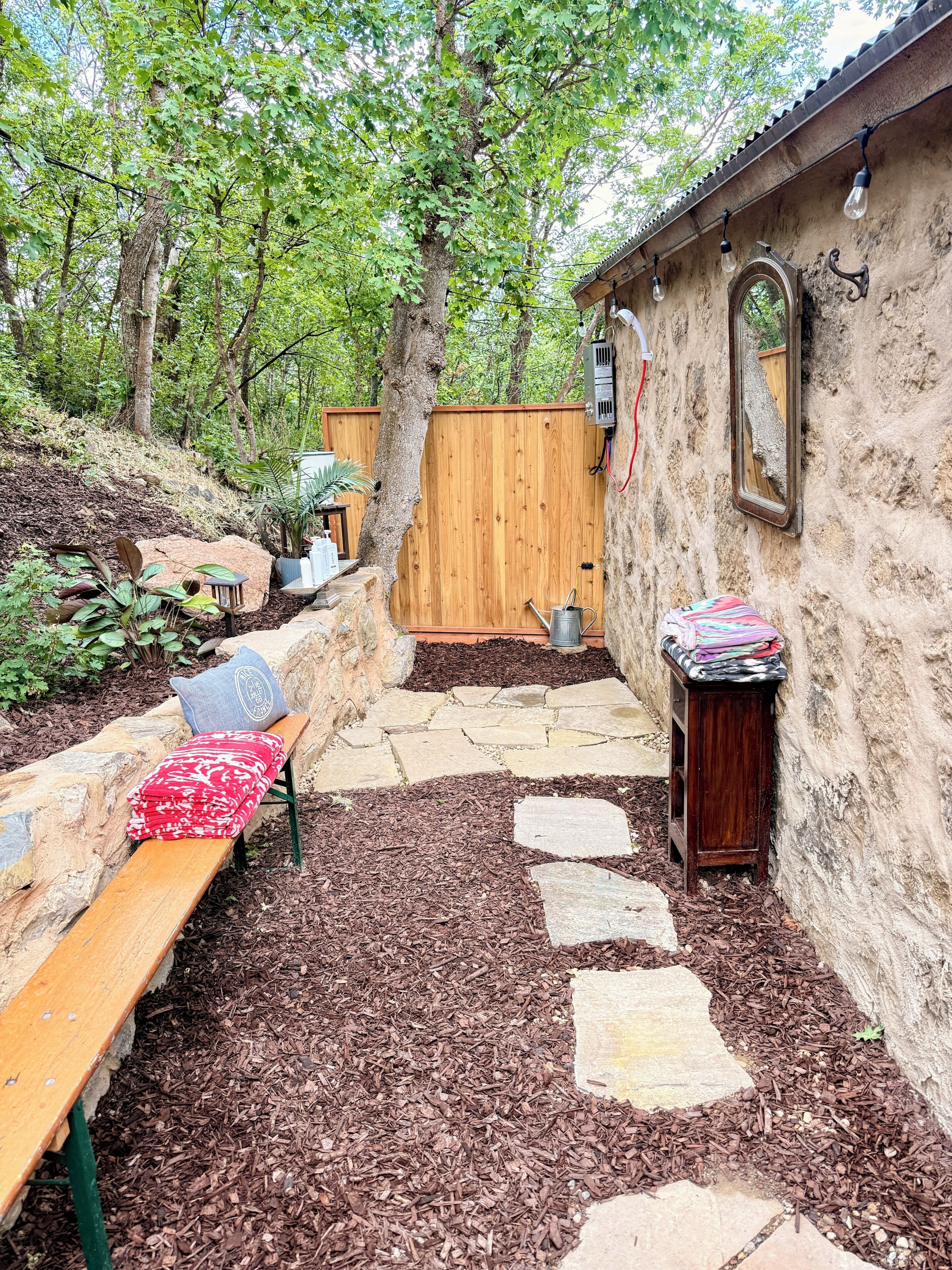 A wooden bench with a red and white blanket, a small table with a colorful cloth, and a stone pathway leading to a wooden fence.
