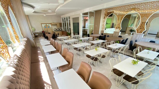 A spacious dining area with multiple tables and chairs, a long cushioned bench along one wall, and decorative elements like mirrors and patterned screens. - Grand Bunda Hotel Syariah Bukittinggi Powered By Archipelago (Bukittinggi)