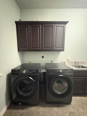 A laundry room with two front-loading washing machines, a dark wooden cabinet above, and a sink with a faucet to the right. - Lakefront Vacation Home: Pool, BBQ, Fire-pit, Theatre, Basketball, Bike, Boat (The Woodlands)