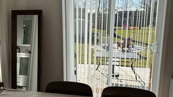 A dining area with a table and chairs, a wooden bowl on the table, and a view through a window with blinds to an outdoor patio with a table and chairs.