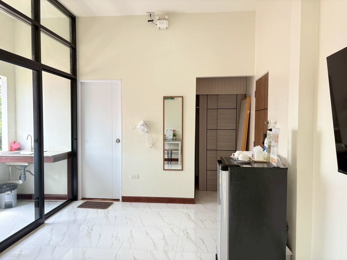 A hotel room with a large sliding glass door leading to a balcony, a white door, a mirror, a small table with toiletries, and a dark-colored cabinet.