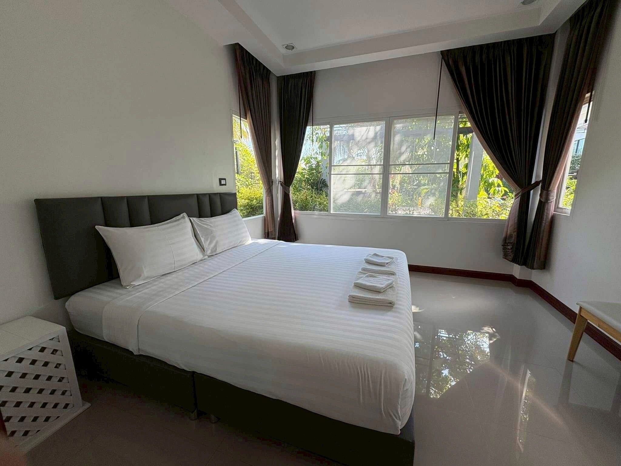 A neatly made bed with white linens and pillows, a dark headboard, and a window with curtains.