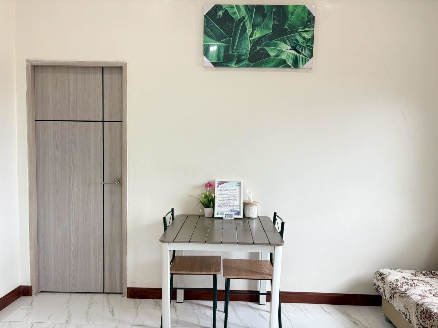 Una habitación con puerta de madera, una pequeña mesa con dos sillas, un cuadro enmarcado en la pared y una cama con cubrecama estampado.