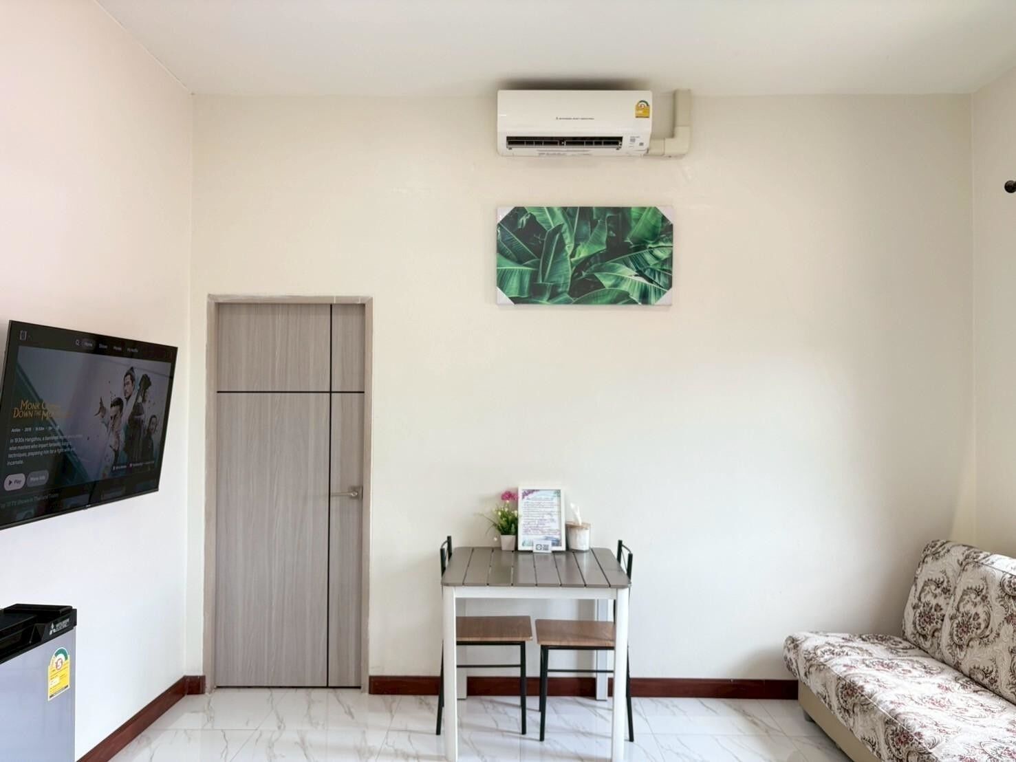 A room with a wooden door, a small table with two chairs, a sofa, an air conditioning unit, and a framed picture on the wall.