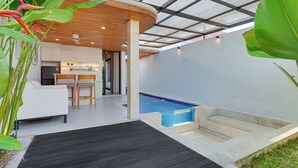 Una cocina moderna con armarios blancos, una zona de comedor con sofá y sillas blancos, techo de madera y una pequeña piscina interior.