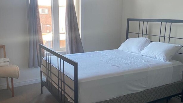 A bedroom with a single bed, a window with curtains, a chair, and a patterned rug. - Divinity Hut (Brampton)