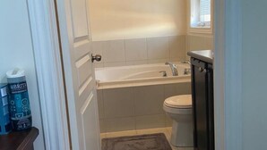 A small bathroom with a bathtub, toilet, and a window. There are two rugs on the floor. - Divinity Hut (Brampton)