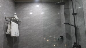 A modern bathroom with a glass-enclosed shower, a towel rack with a white towel, and a small shelf with toiletries. - Twin Sky Suite – Spacious, Stylish, Serene (Parañaque)