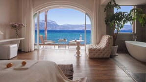 Ένα σύγχρονο υπνοδωμάτιο με ένα μεγάλο παράθυρο, μια μπανιέρα και έναν χώρο καθιστικού με θέα στη θάλασσα.