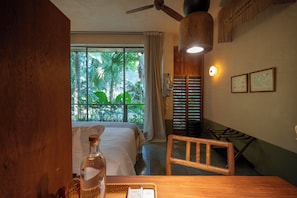 A hotel room with a bed, a wooden chair, a table with a bottle and menu, a ceiling fan, and a view of tropical plants through the window. - Casa Querida — A Private Oasis in the Heart of Mérida (Mérida)