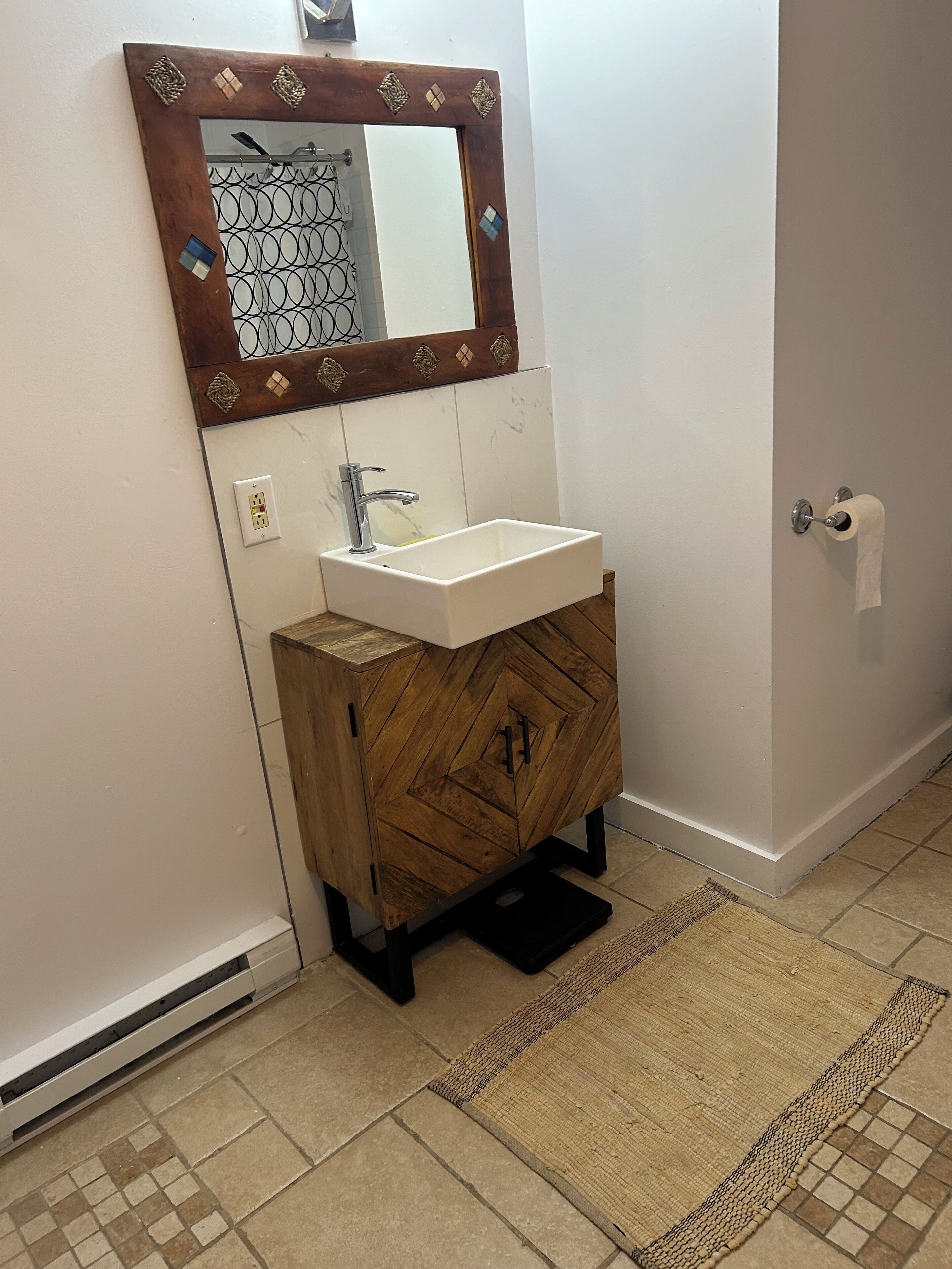 Une salle de bain avec un meuble en bois, un lavabo blanc, un miroir avec un cadre décoratif et un porte-rouleau de papier toilette.