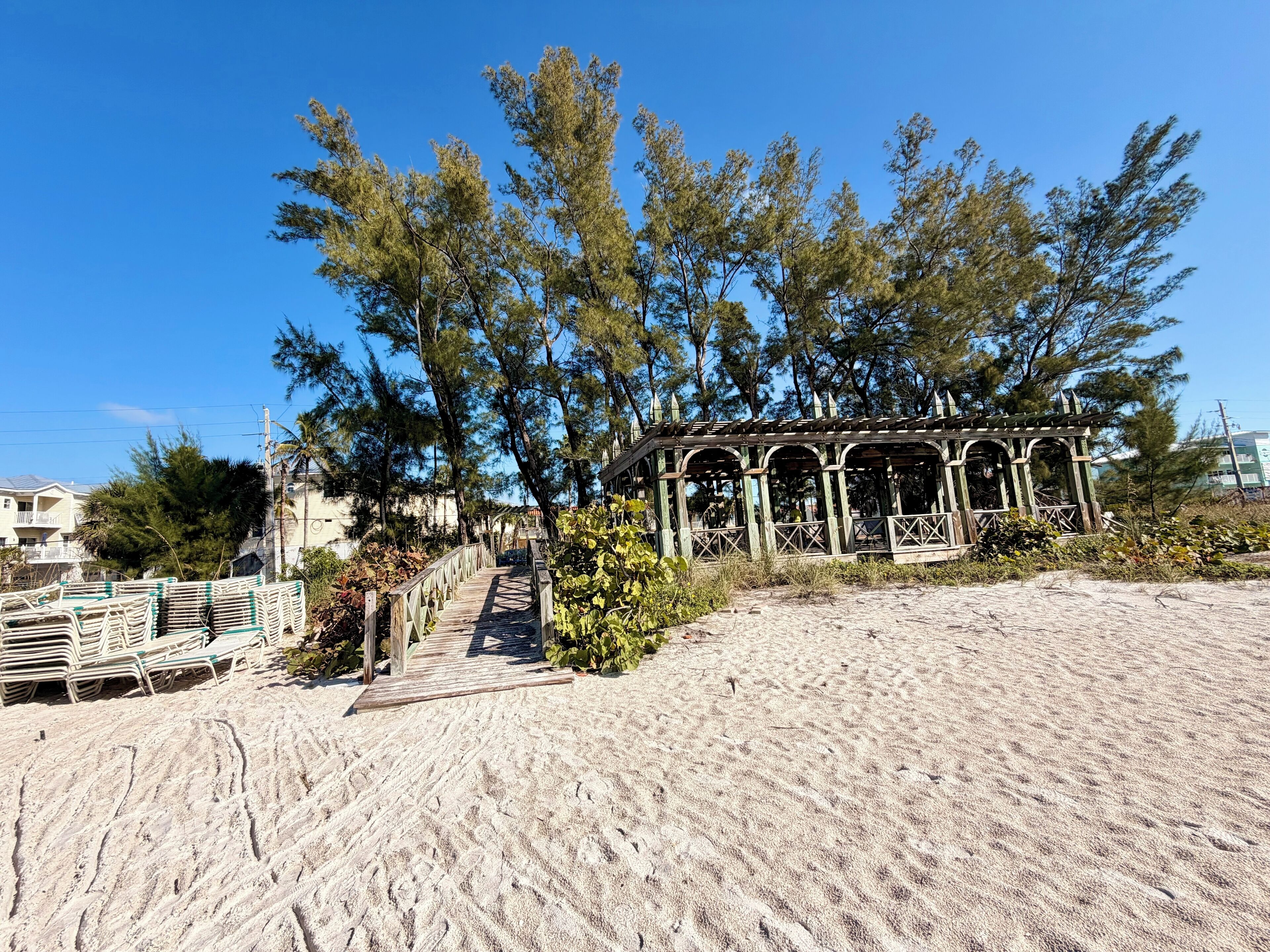 A wooden gazebo on a sandy beach with a staircase leading up to it, surrounded by tall trees.