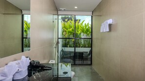 A modern bathroom with a glass shower enclosure, a seating area with chairs, and a large mirror reflecting the room. - Adva Garden Village (Canggu)
