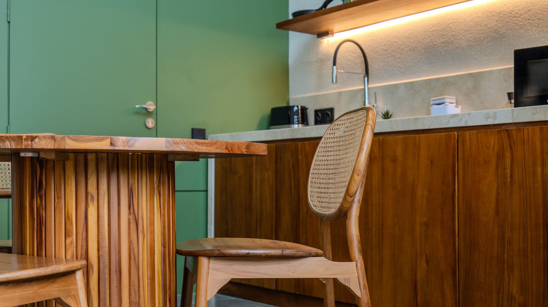 A wooden dining table with a woven chair, a green door, and a kitchen counter with a sink and appliances.
