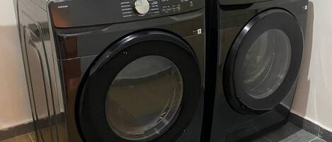 Deux machines à laver à chargement frontal de couleur noire, placées dans un coin, avec un sol carrelé et une prise électrique fixée au mur.