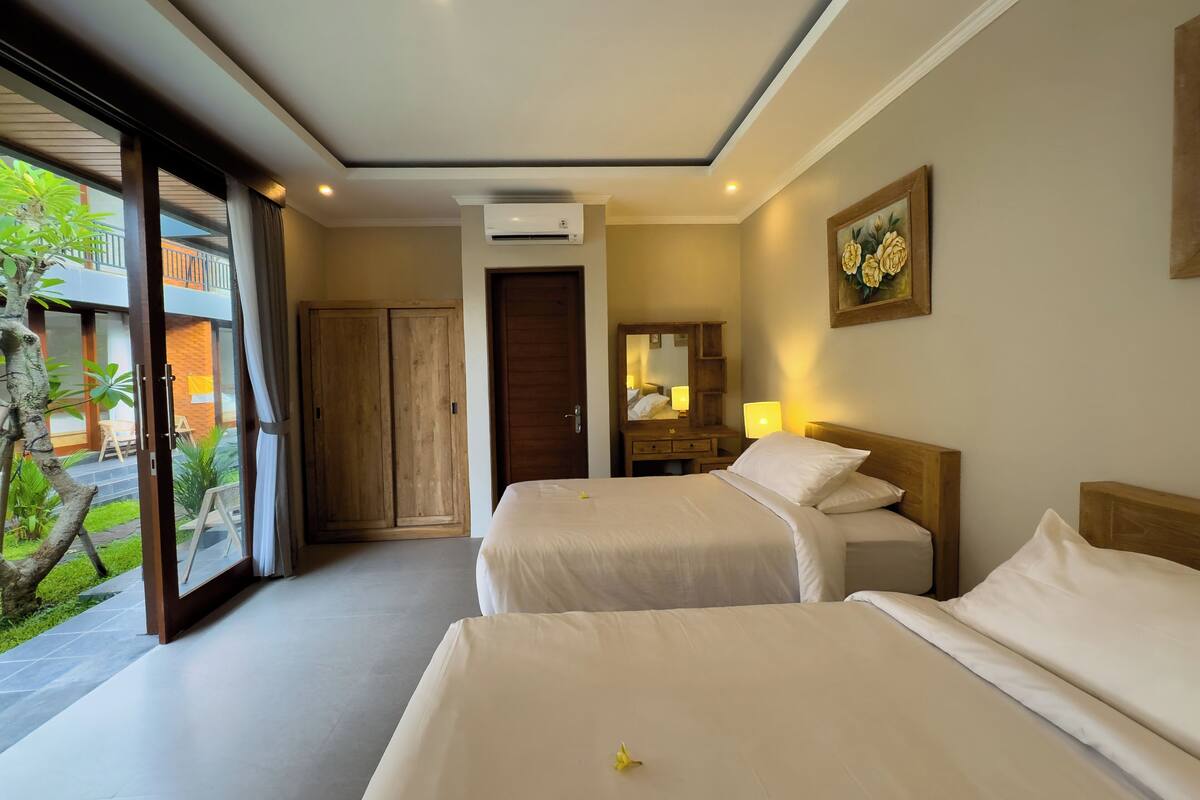 Ein Hotelzimmer mit zwei Betten, einem Holzschrank, einem Badezimmer inklusive Spiegel und Blick auf einen Balkon mit Pflanzen.