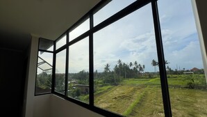 Ein Zimmer mit großen Fenstern, das einen Blick auf eine ländliche Landschaft mit Feldern und Palmen bietet.