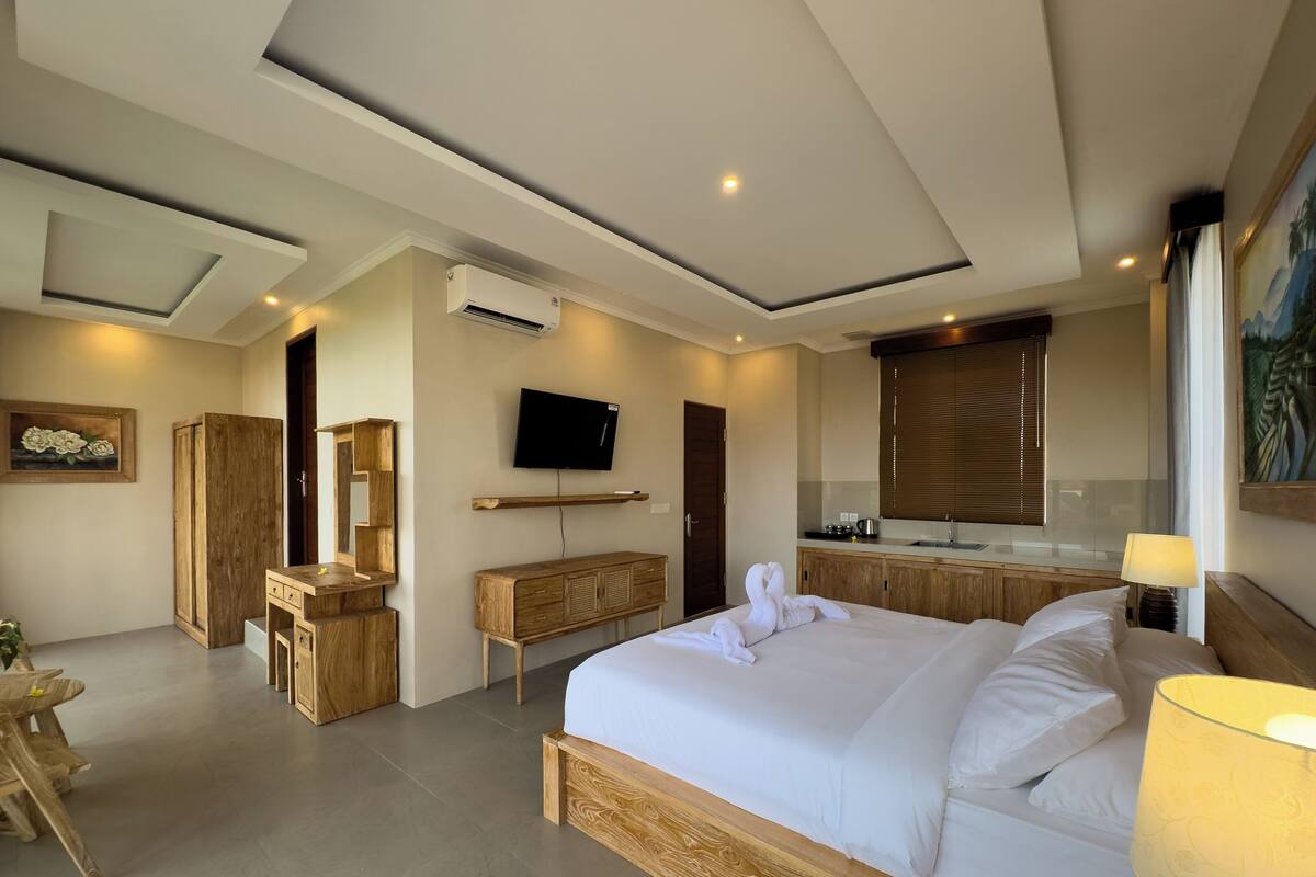 Ein modernes Hotelzimmer mit einem großen Bett, Holzmöbeln, einem Flachbildfernseher, einer kleinen Küchenzeile und einem großen Fenster mit Jalousien.