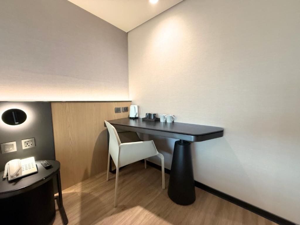 Quarto moderno com escrivaninha de madeira escura, uma cadeira branca, uma mesinha redonda com telefone e um cesto de lixo preto.