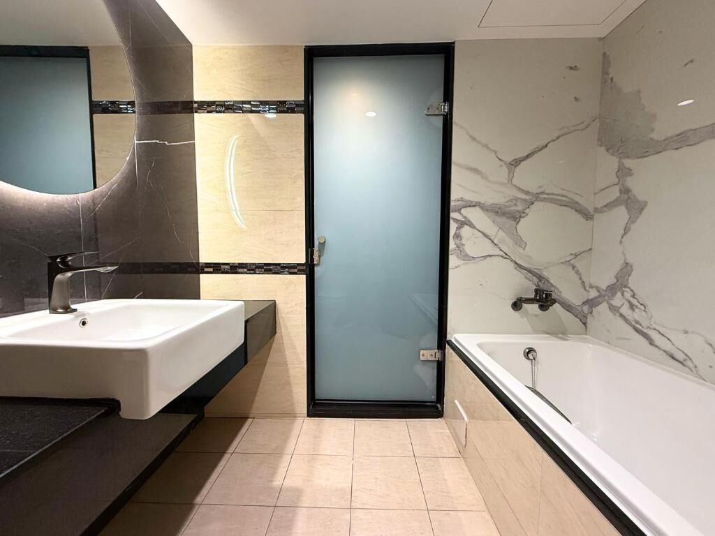 A modern bathroom with a large mirror, a white sink, a bathtub, and a frosted glass door.