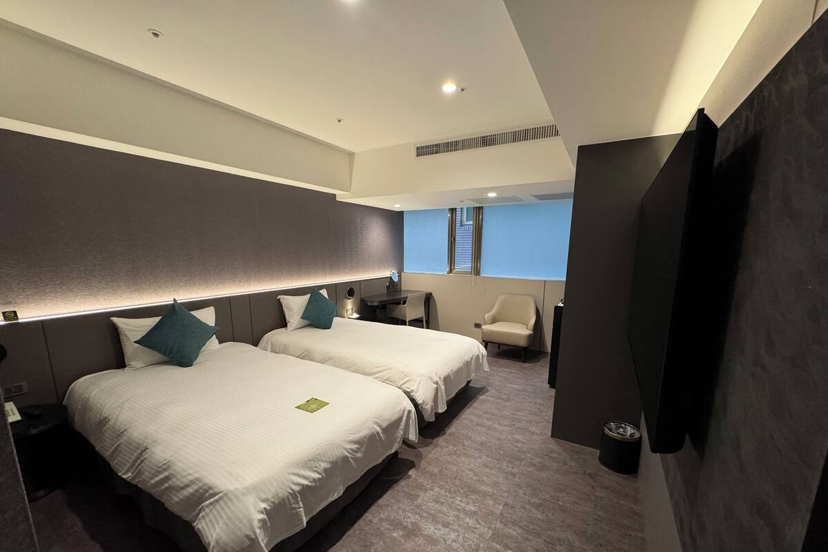 A hotel room with two beds, a desk, a chair, a TV, and a window.