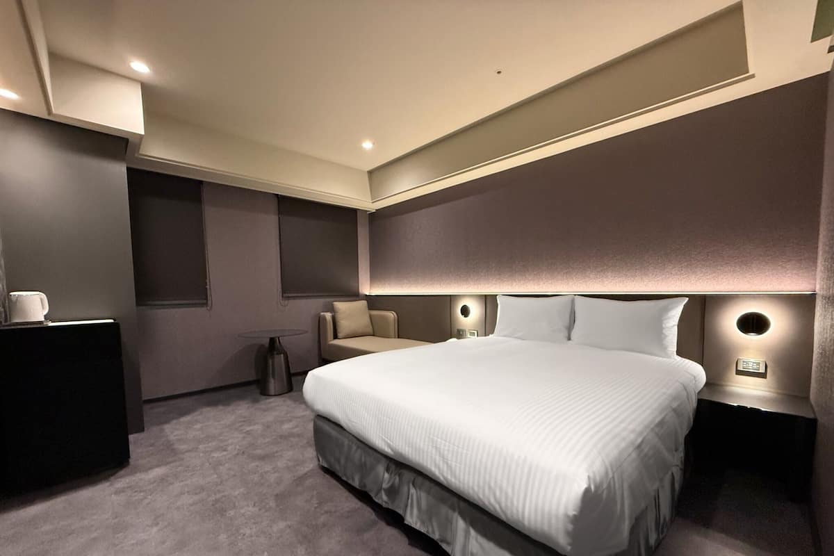 A modern hotel room with a large bed, a small seating area, and a desk with a cup.