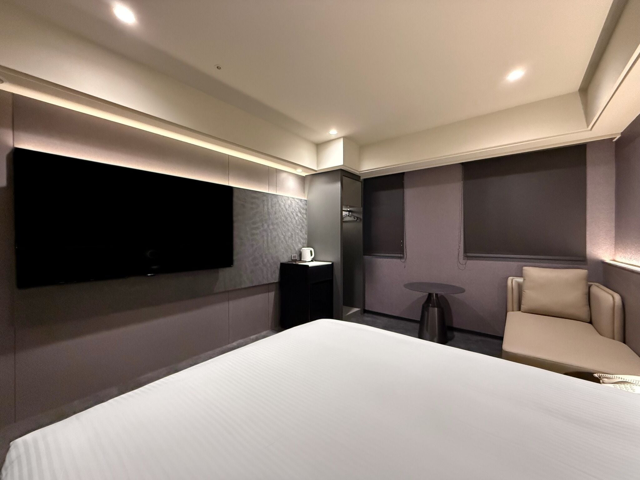 Una habitación de hotel moderna con una cama grande, un televisor de pantalla plana en la pared, una mesita redonda y un sillón cómodo.
