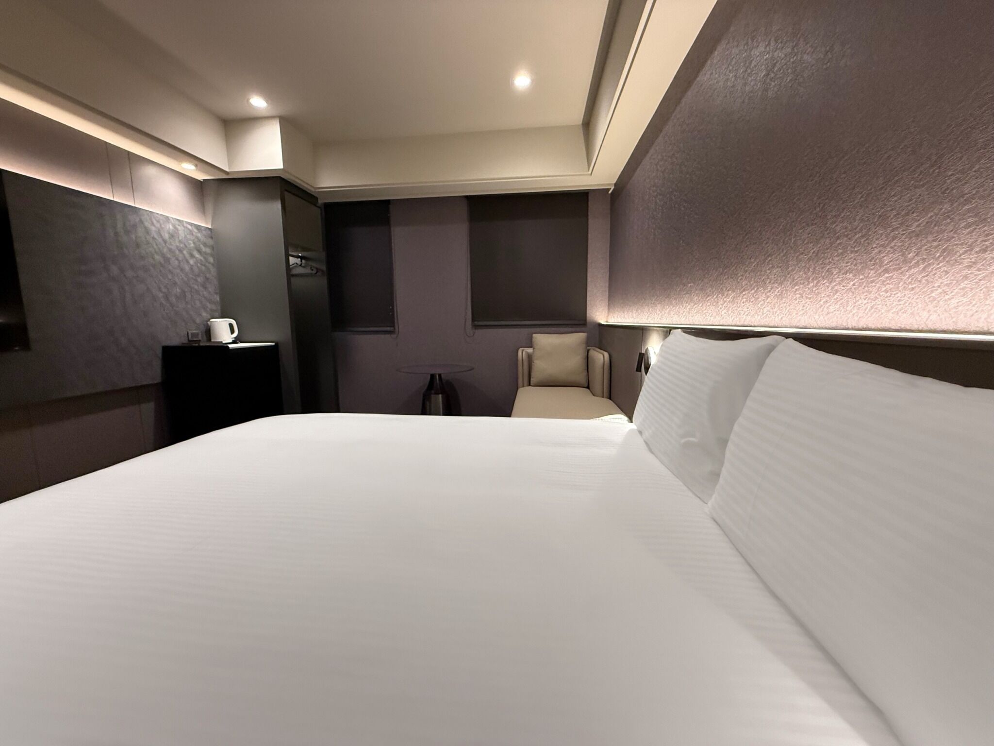 A modern hotel room with a large bed, a bedside table, a chair, and a lamp.