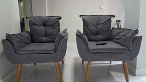 Zwei graue Sessel mit Holzbeinen, ein schwarzer Standventilator und eine Fernbedienung auf einem der Sessel.
