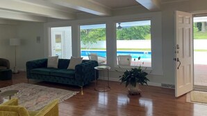 A spacious living room with a green velvet sofa, a small round table, and a large window overlooking a swimming pool. - Spacious Home with Heated Swimming Pool + Hot Tub + Event Room + SLEEPS 23 (Owensboro)