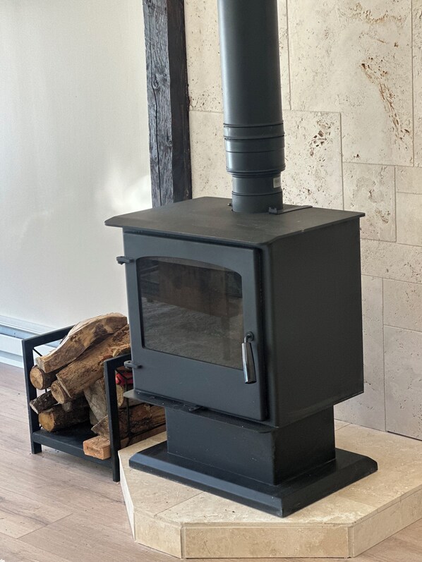 A black wood-burning stove with a glass door, positioned on a tiled floor next to a stack of firewood. - Modern Cozy barn by the creek (Evergreen)