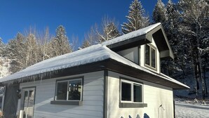 Una cabaña de dos pisos con techo nevado, un porche con sillas Adirondack y cimientos de piedra.
