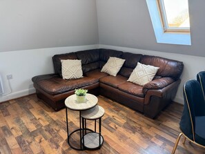 A corner living room with a brown leather sectional sofa, two decorative pillows, a small round table with a plant, and a window with a view of trees. - Guildford Court Apartment Colchester (Colchester)