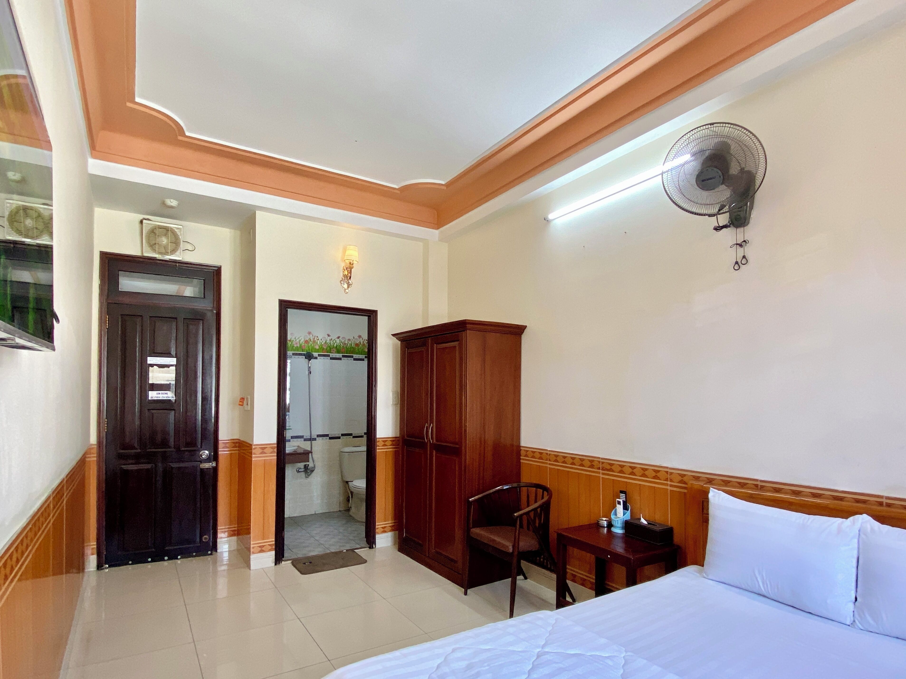 Hotelkamer met een bed, een houten kledingkast, een stoel, een nachtkastje met een lamp, een wandventilator, en een zichtbare badkamer door een open deur.