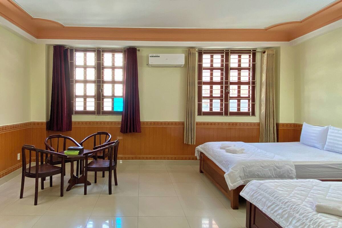 A hotel room with two beds, a wooden table with chairs, and two windows with curtains.