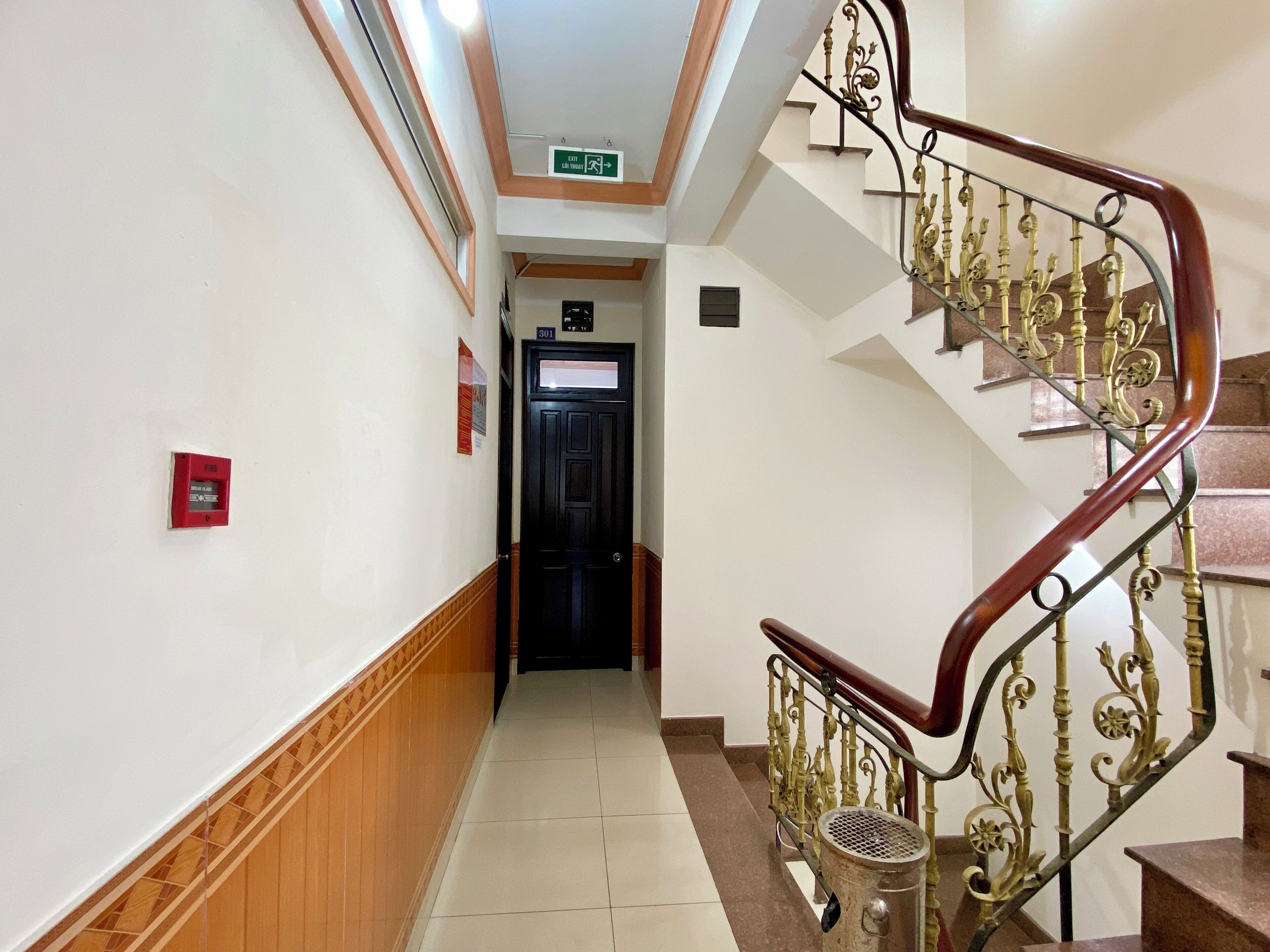 Un couloir d’hôtel avec un plancher en bois, une porte noire portant un numéro et un escalier avec des rampes en métal décoratives.