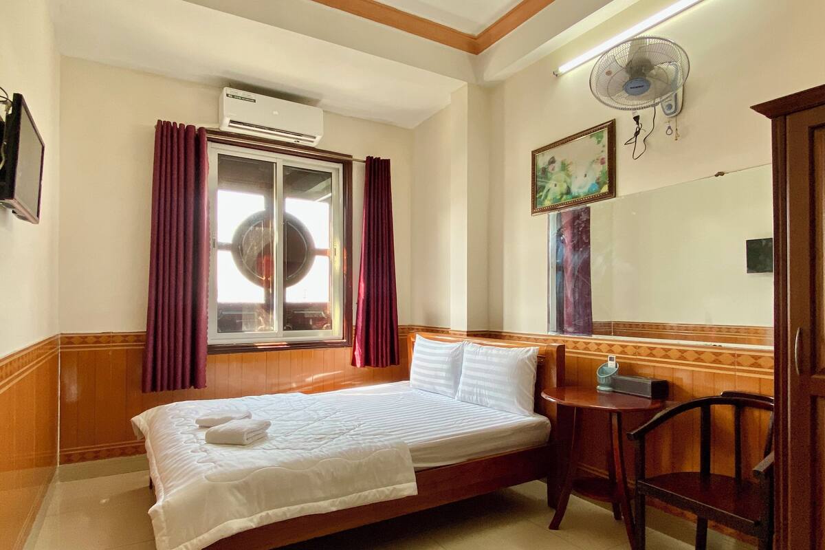 A hotel room with a bed, a desk, a chair, an air conditioner, a window with curtains, and a wall-mounted fan.