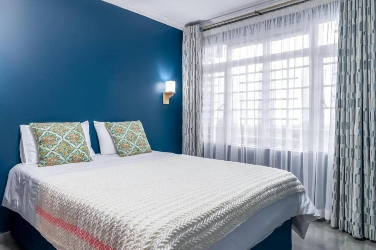 Une chambre d’hôtel avec un grand lit, deux coussins à motifs, une couverture blanche et une fenêtre ornée de rideaux légers.