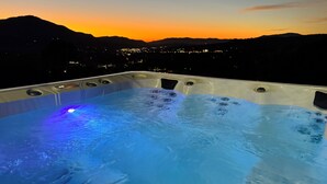 Un bain à remous avec jets, entouré d’un paysage montagneux et illuminé par les lumières de la ville en arrière-plan.
