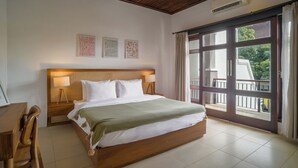 Ein Schlafzimmer mit einem großen Bett, einem Holzkopfstück, zwei Nachttischlampen, einem Schreibtisch mit Stuhl und einem großen Fenster mit Blick auf Grünflächen.