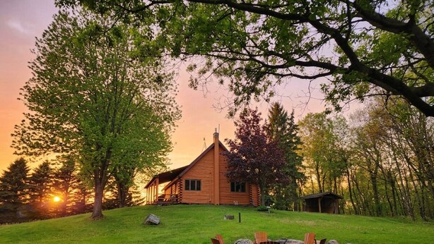 Ein Blockhaus mit Feuerstelle und Adirondack-Stühlen auf einer grünen Wiese.