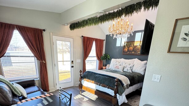 A bedroom with a bed, a table, and a chair. There is a window with curtains, a chandelier, and a plant. - Cozy cottage with fresh baked goods (Colorado City)