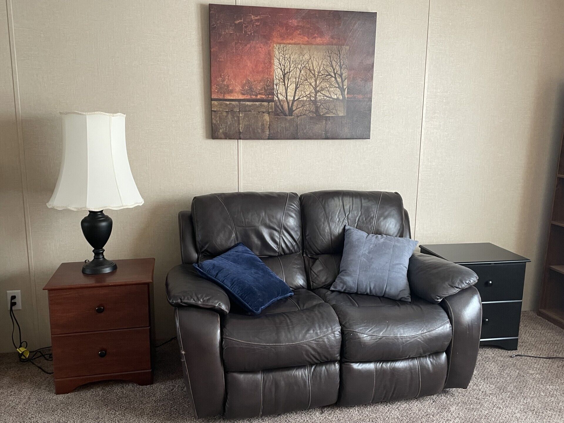 A leather recliner with two cushions, a small wooden nightstand with a lamp, and a painting on the wall.
