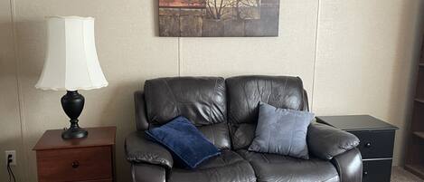 A leather recliner with two cushions, a small wooden nightstand with a lamp, and a painting on the wall.